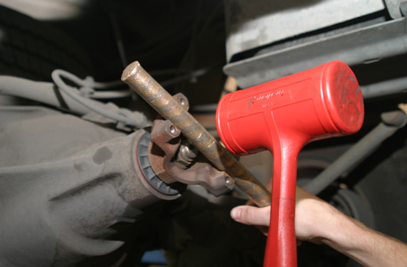 Tight Yoke Installation - In many cases when installing new yokes on Ford 10.25”and 10.5” as well as some other differentials, it can be difficult to get the yoke over the pinion splines. The fact is, many pinion splines have a slight “twist” to them (which controls rotational “slop”), but the Ford 10.25” & 10.5” pinion splines have a slightly tighter fit than most. As a result, they make it difficult to get a new yoke over the pinion splines, and it’s an even bigger challenge when installing a new yoke over a new pinion gear.

The solution? There really isn’t one, you’ve just got to use good old-fashioned force. Generally, it’s a two person job. One technician holds the pinion in the backside of the housing, while the other cradles a brass punch in the yoke and use a dead blow mallet to drive the yoke into place (See Pic). Keep striking the yoke until the pinion nut threads are exposed enough to get a few turns of the pinion nut on the pinion, then use an impact gun to get the yoke seated. (Be careful to make sure to have enough pinion thread showing to ensure you won’t strip the threads off the pinion.) It sounds rough, we know…but it’s the only way to get the job done.